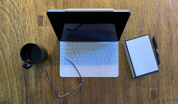 A laptop on a table, with a coffee mug, glasses, notebook and pen