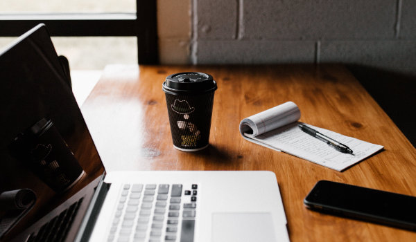 A laptop, cup of coffee, and a pen and notepad on a desk